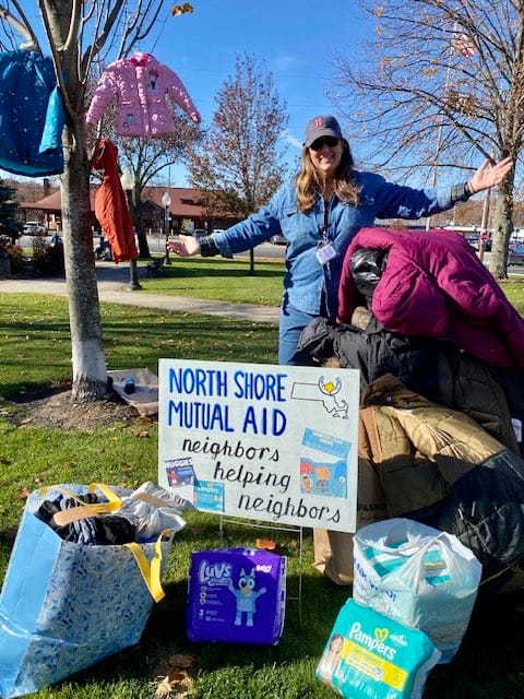 Photo of a white woman wearing a blue coat and Red Sox baseball cap standing around piles of donated coats and diapers. She has her arm outstretched. In the foreground is a sign that reads, "North Shore Mutual Aid: Neighbors Helping Neighbors